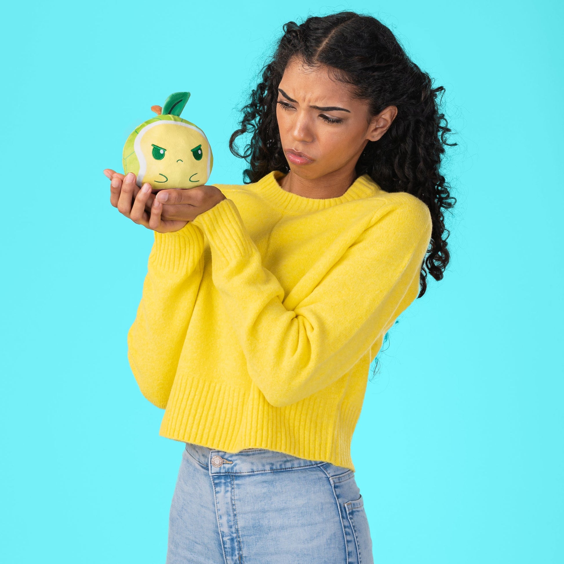 A woman wearing a yellow sweater, holding a TeeTurtle Plushiverse Turtley Delicious 4” Reversible Plushie.