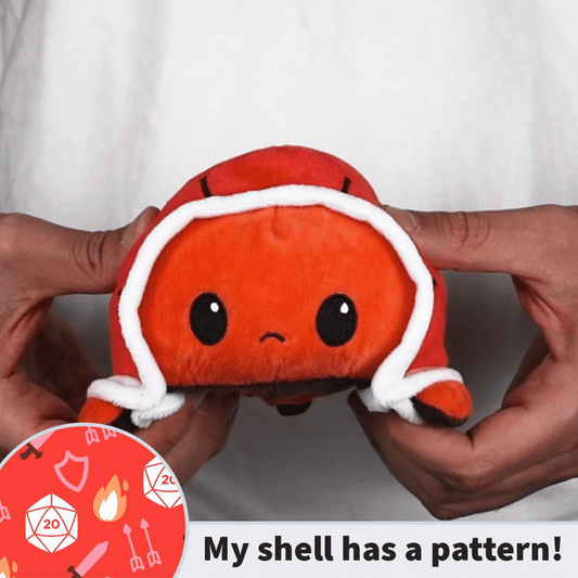A close-up of someone holding an adorable red TeeTurtle Reversible Turtle Plushie, featuring a sad expression and adorned with a shell trimmed in white. The text overlay reads,