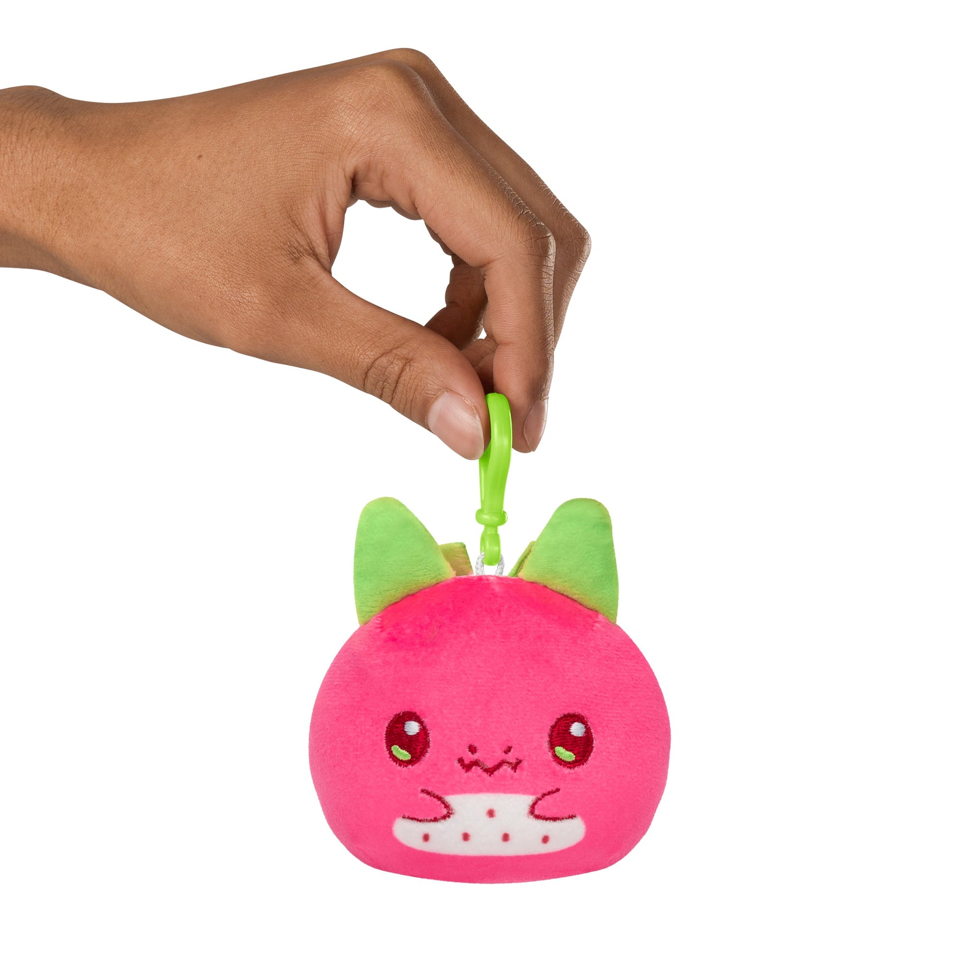 A hand attaching a Plushiverse Dragonfruit Reversible Plushie Keychain to a pink plush toy with cat-like features and green ears, isolated on a white background.