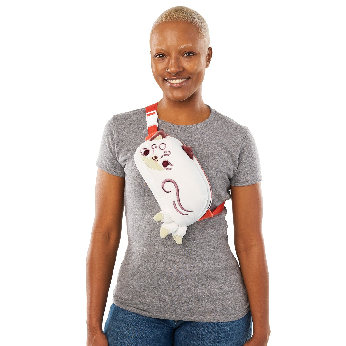 A smiling woman with a shaved head wearing a gray t-shirt and jeans, carrying a whimsical Plushiverse Magical Kitsune Fanny Pack from the TeeTurtle collection over her shoulder.